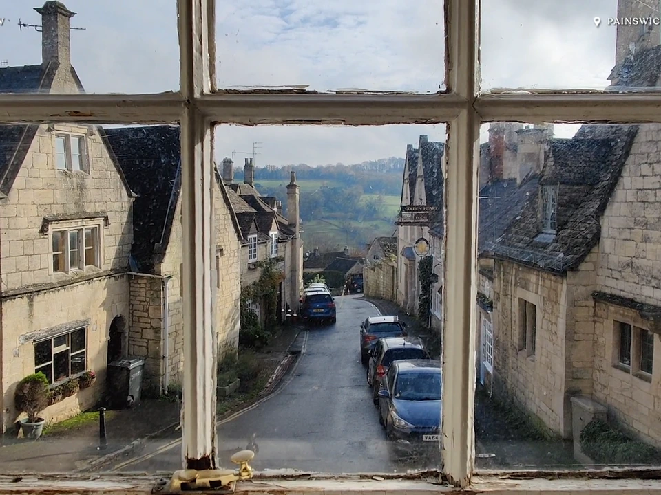 View through an old window overlooking a narrow village street lined with stone buildings and parked cars, with green countryside visible in the distance.