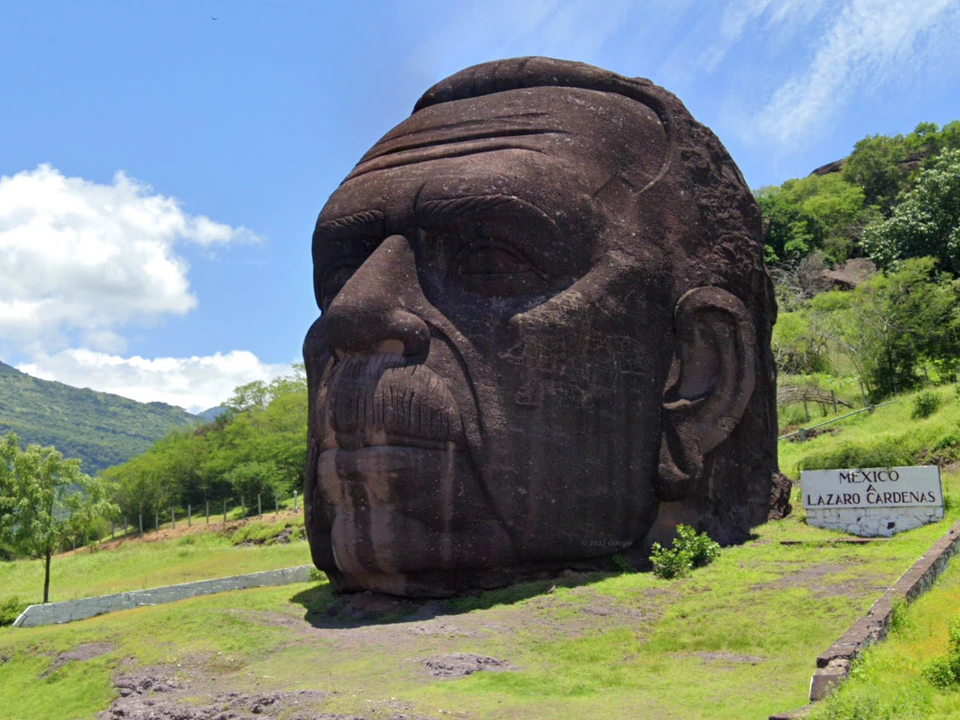 Large carved stone head sculpture outdoors on a grassy hillside beneath a bright blue sky.