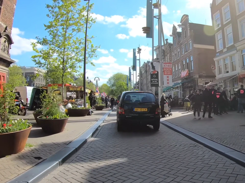 Street-level view from a car driving through a sunny city center, with another car ahead, pedestrians along the sidewalks, shops and historic buildings, and trees lining the street.